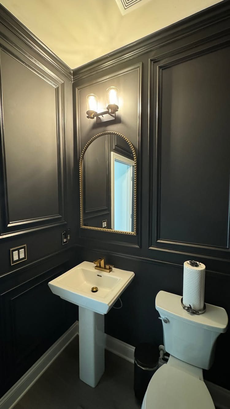 Dark charcoal paneled walls with decorative molding surround a white pedestal sink and gold mirror.