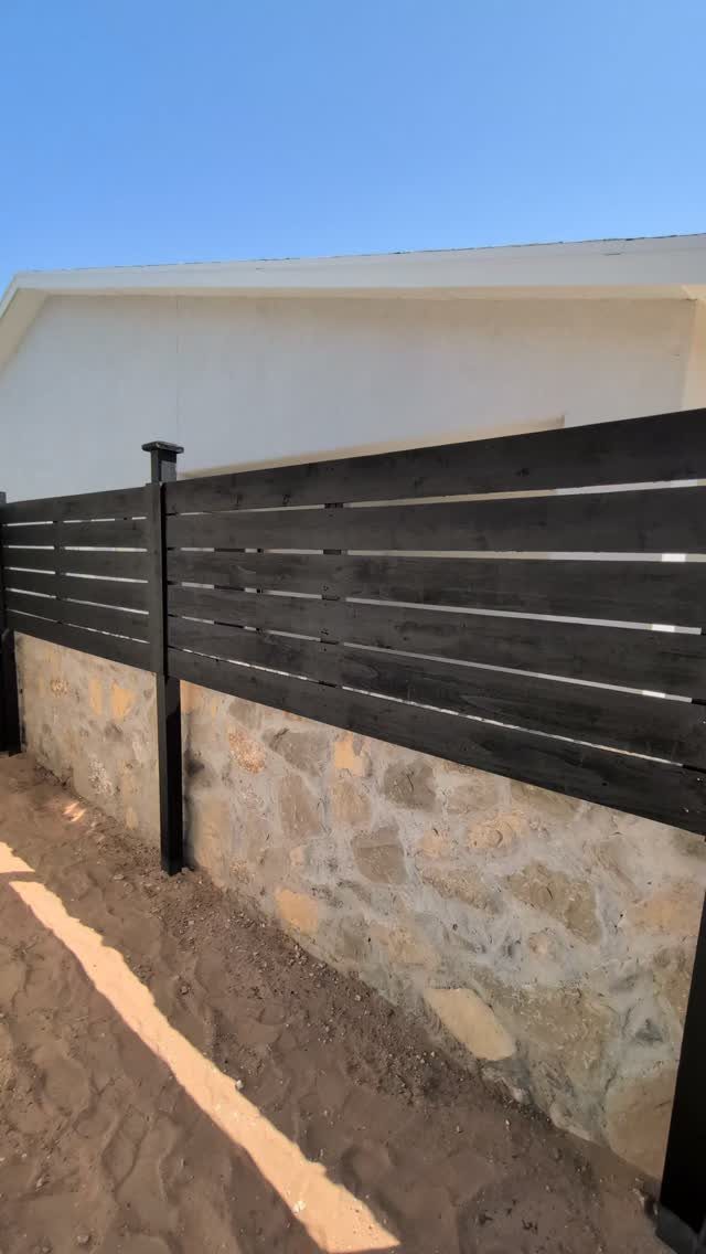 Black horizontal wood slat fence installed on a stone retaining wall against a blue sky.