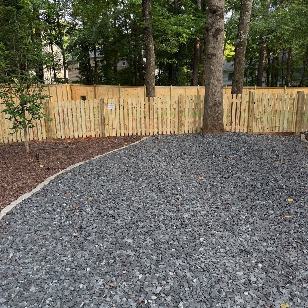 A light wood picket fence borders a yard filled with grey gravel and dark mulch.