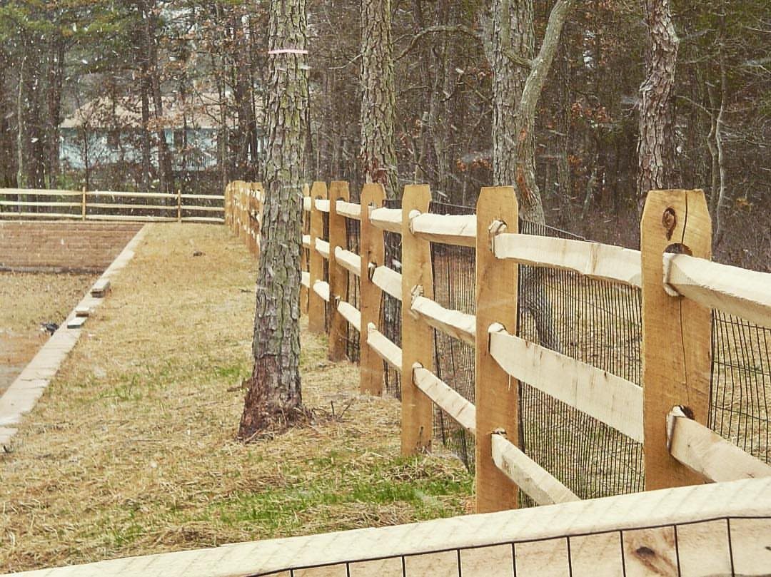 A wooden split rail fence with wire mesh runs along a grassy yard near a forest.