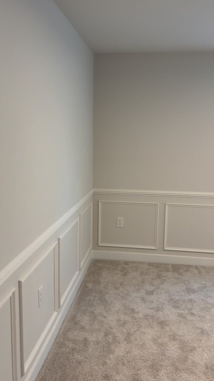 White wainscoting paneling with a chair rail installed on light grey walls in a room with beige carpet.