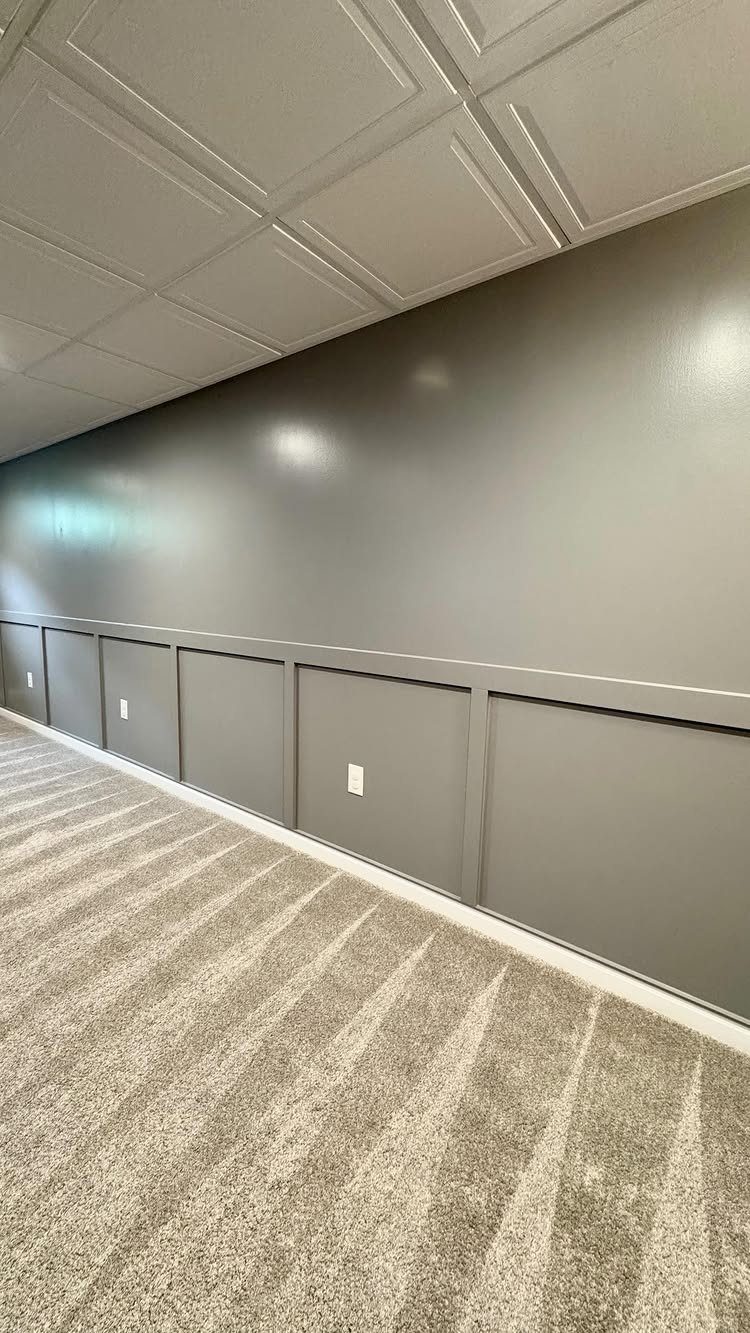 Grey paneled wall with a horizontal chair rail and vertical battens above beige carpeting.