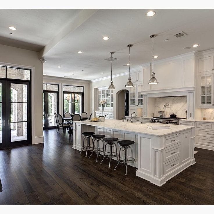Dark hardwood flooring in a large, modern white kitchen with a marble island and black stools.