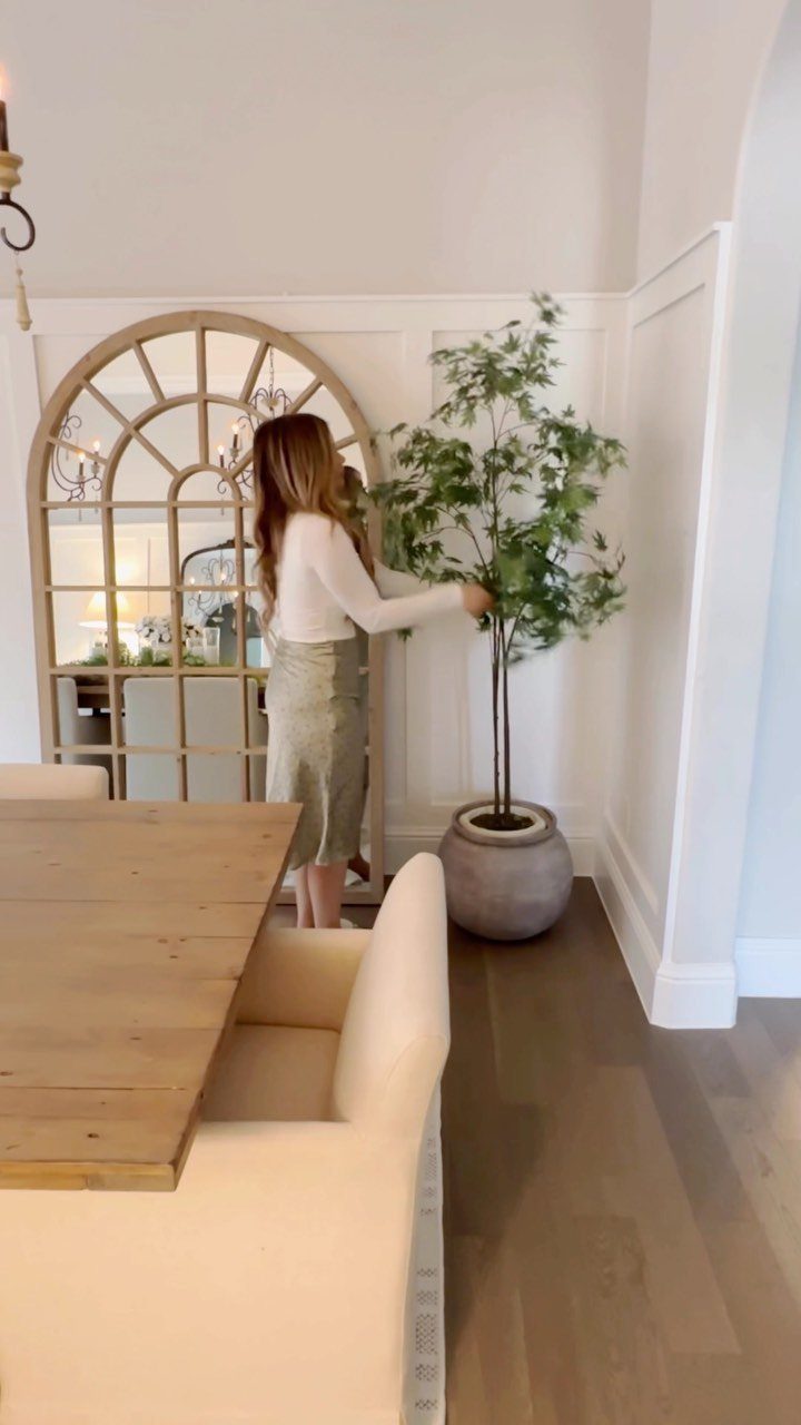 A woman standing next to a large potted plant in a dining room with an arched mirror.