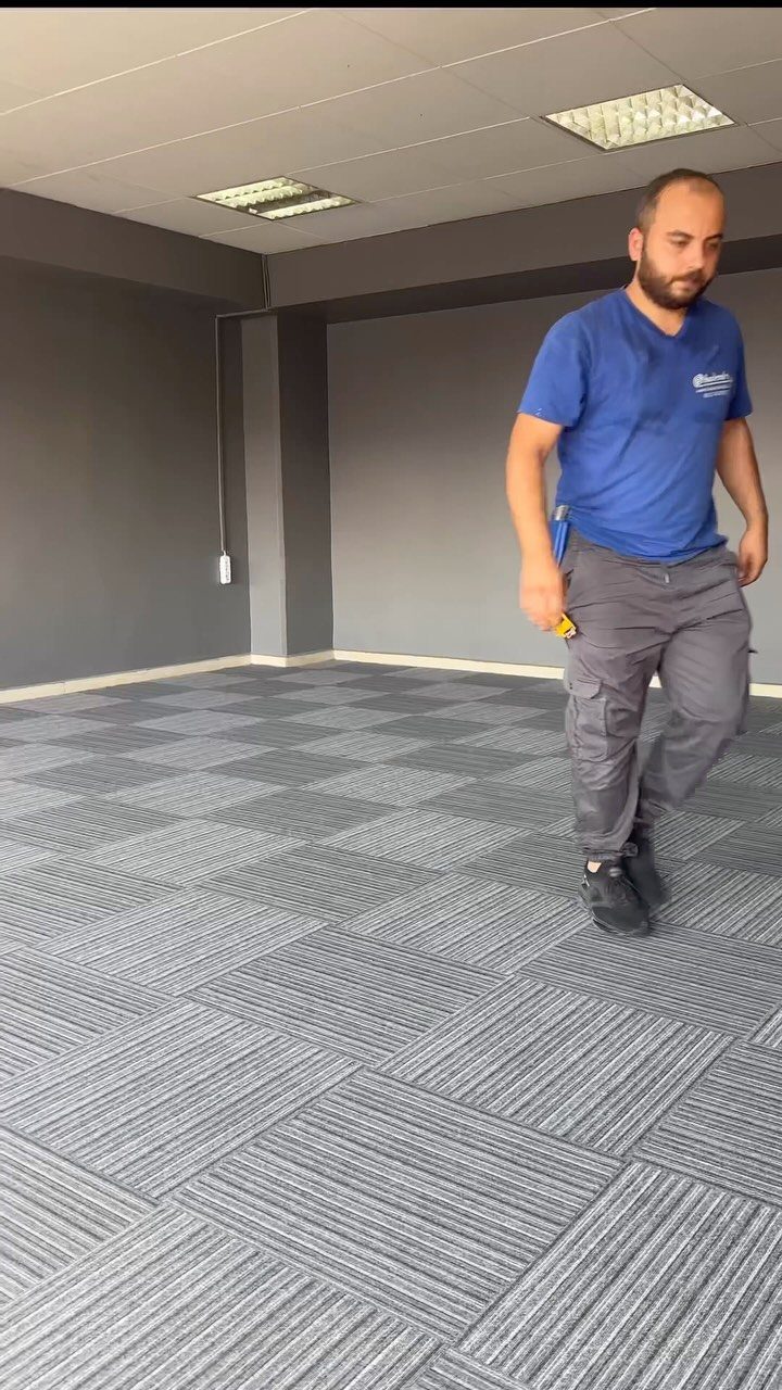 A man walking across a room with grey patterned carpet tiles and dark walls.