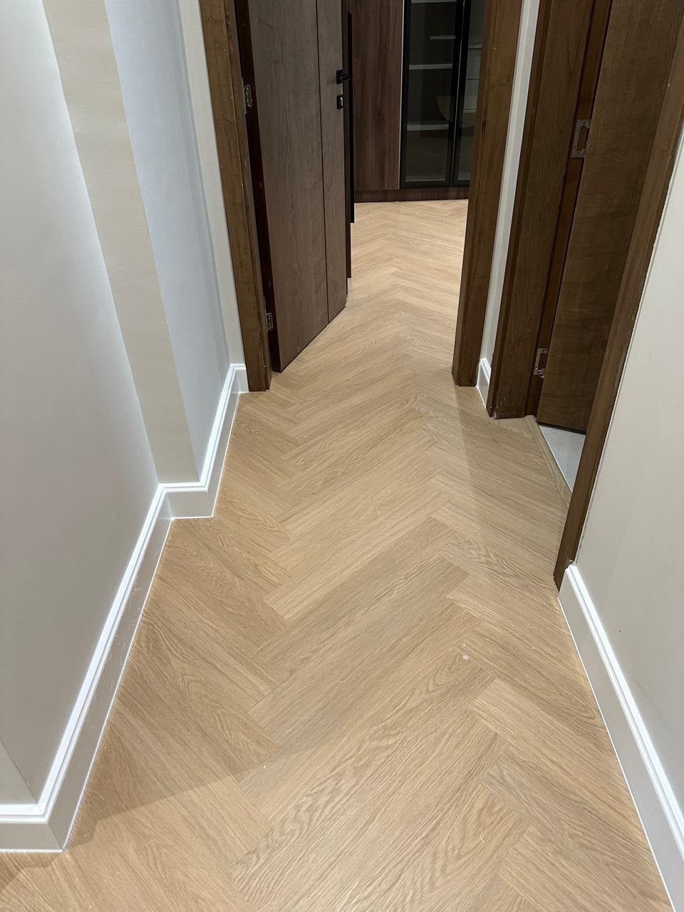 Light wood herringbone patterned flooring running through a hallway with white baseboards.