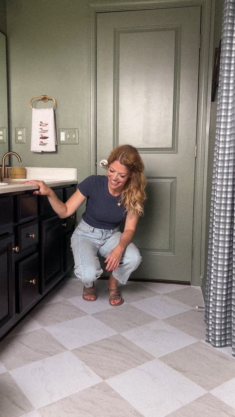 A smiling woman crouches on a checkered grey and white tiled bathroom floor.