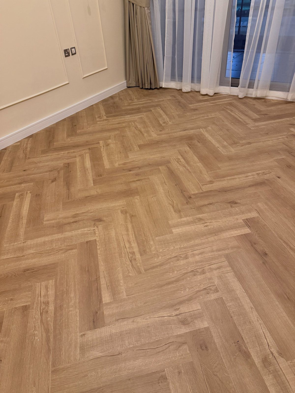 Flooring Trends 2026: 10 Ideas for Warmth, Texture, and Contrast 4 Light wood planks arranged in a herringbone pattern on a floor next to white curtains.