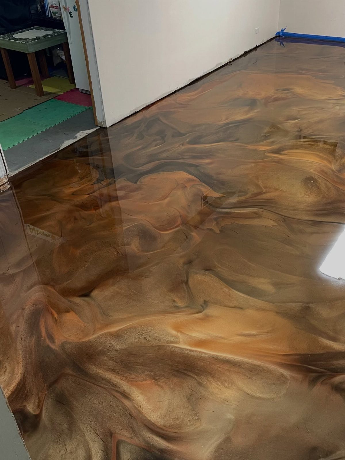 Flooring Trends 2026: 10 Ideas for Warmth, Texture, and Contrast 8 Swirling metallic gold and brown epoxy resin flooring with a high-gloss reflective finish in a room.