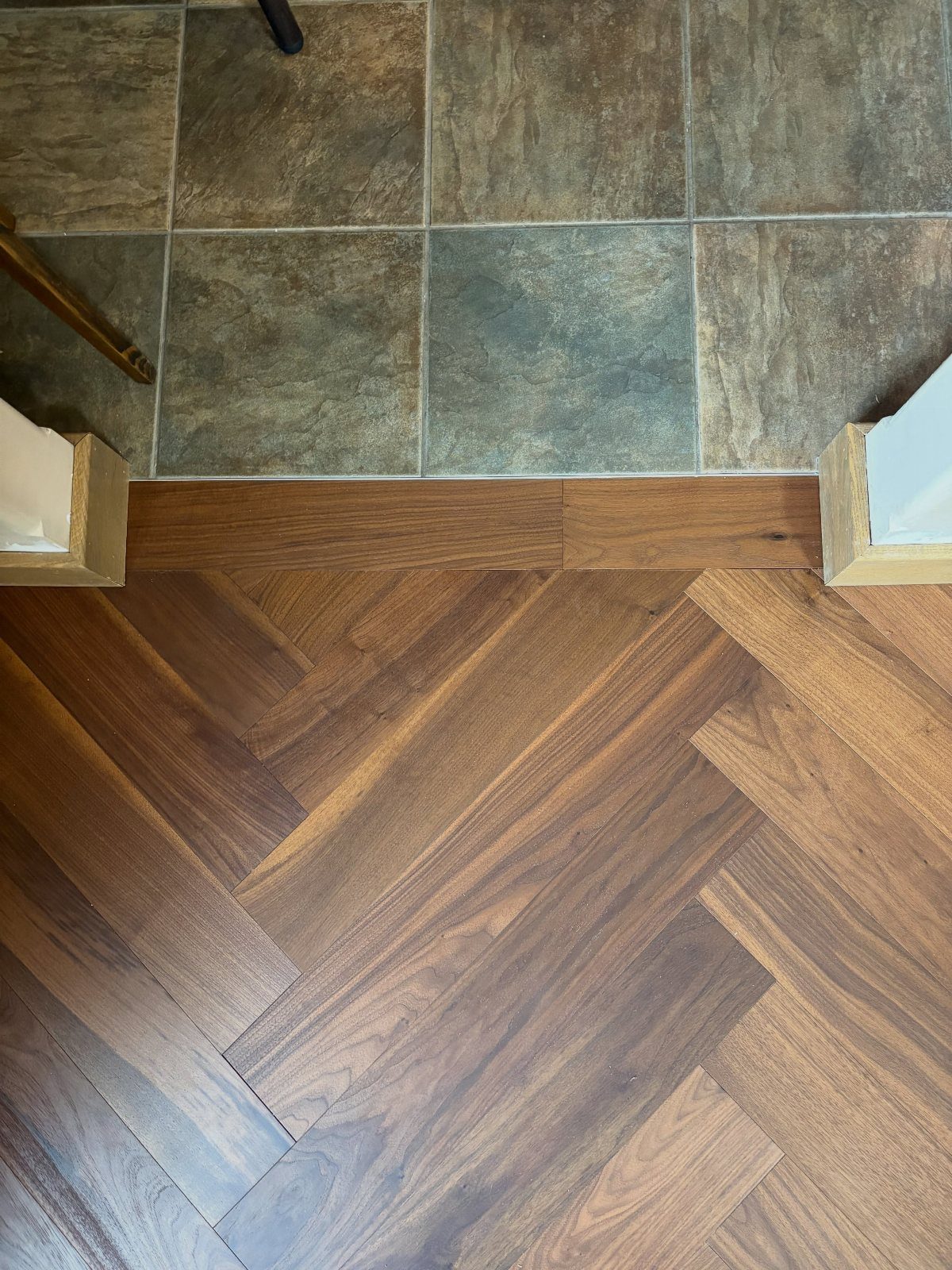 Flooring Trends 2026: 10 Ideas for Warmth, Texture, and Contrast 11 Dark wood herringbone flooring meeting large earth-toned stone tiles at a wooden transition strip.