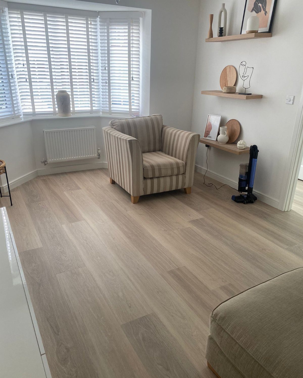 Flooring Trends 2026: 10 Ideas for Warmth, Texture, and Contrast 2 Light neutral oak effect flooring in a bright living room with a striped armchair.