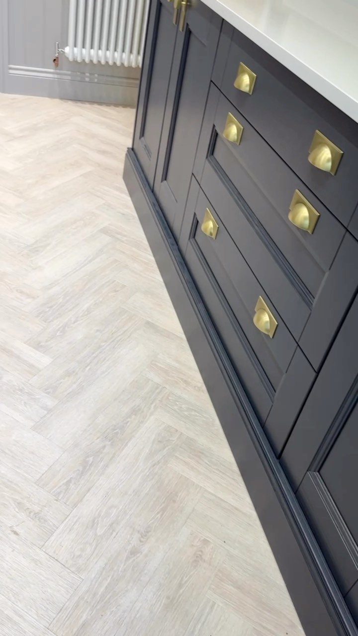 Light grey herringbone patterned flooring installed next to dark grey kitchen cabinets with gold handles.