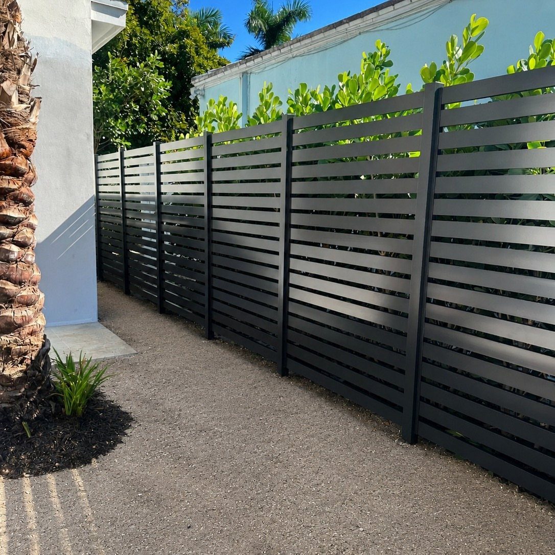 Horizontal Slat Fence: 8 Designs for Modern Backyards 6 Black horizontal slat fence running along a gravel walkway next to a white building and green plants.