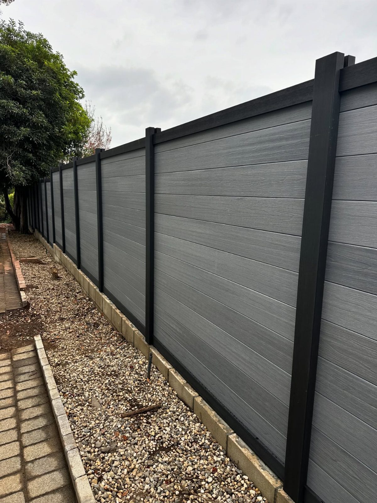 Horizontal Slat Fence: 8 Designs for Modern Backyards 2 Grey composite horizontal slat fence with black posts running alongside a gravel and brick path.
