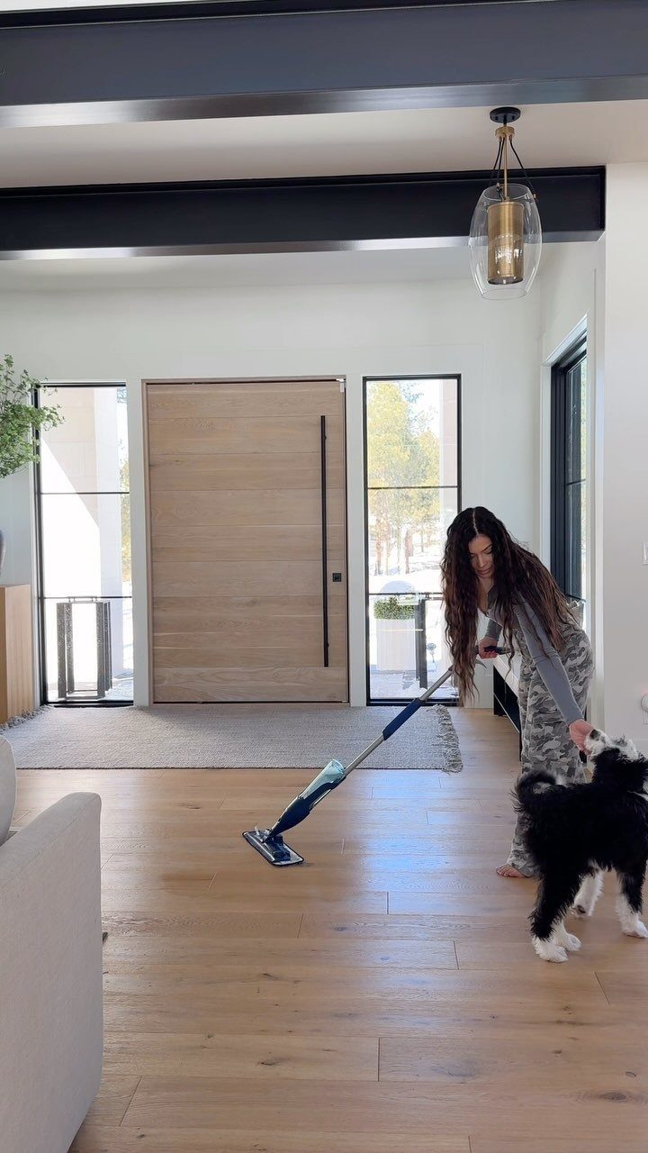 A person mopping light oak wooden floors in a bright, modern living space with a dog.