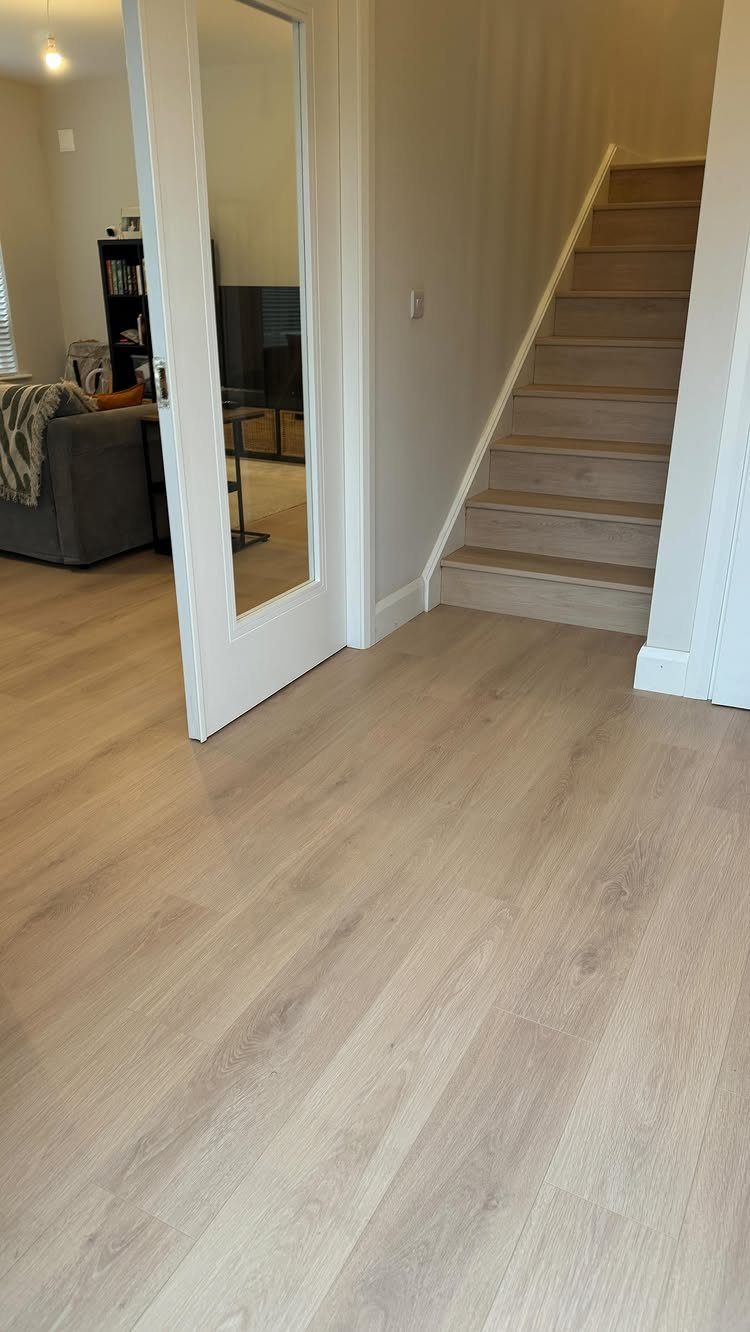 Light wood laminate flooring extending through a hallway towards a staircase and a mirrored door.