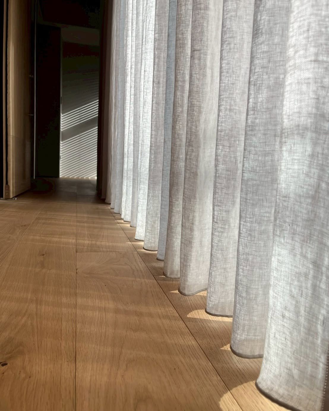 Light wooden floorboards running alongside long, white linen curtains in a sunlit room.