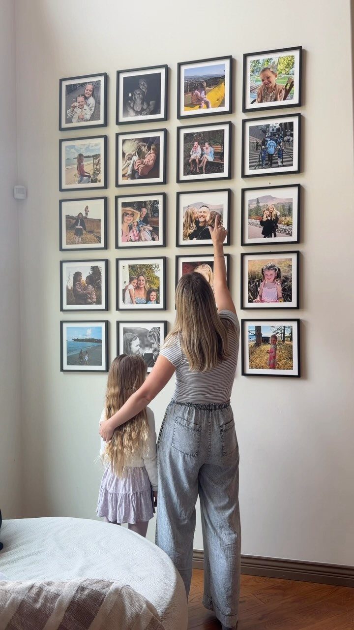 A woman and child arranging a large collection of framed family photos on a white wall.