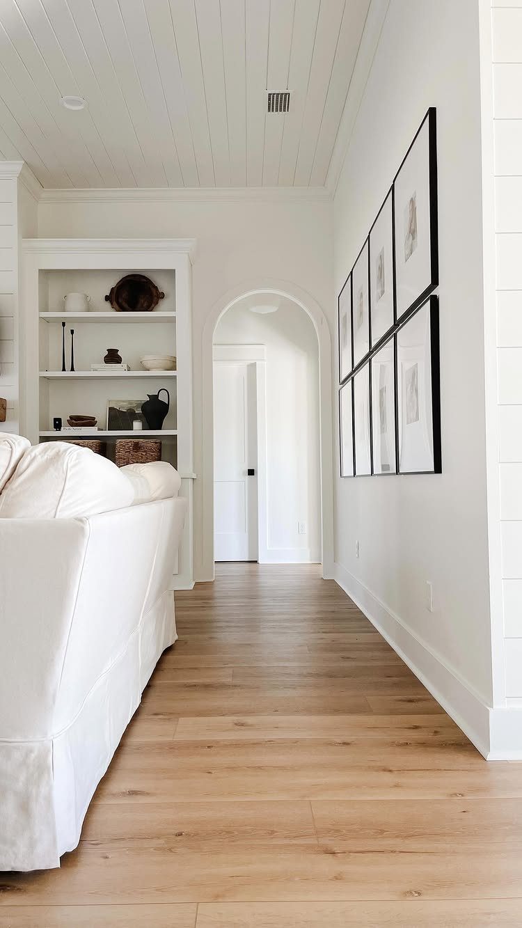 Light wood-plank flooring leading through a bright living room toward an arched white doorway.