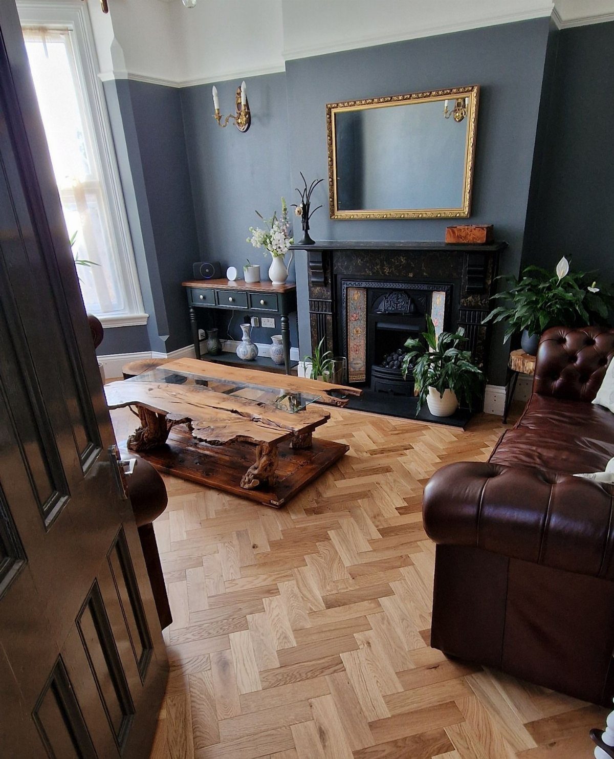 Light wood herringbone flooring in a living room with a leather sofa and dark fireplace.