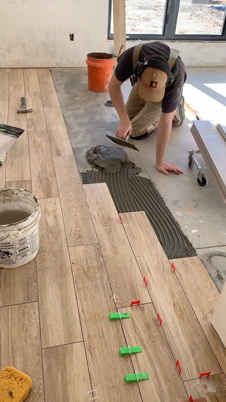 A worker applies mortar to a floor while installing light brown wood-look tiles.