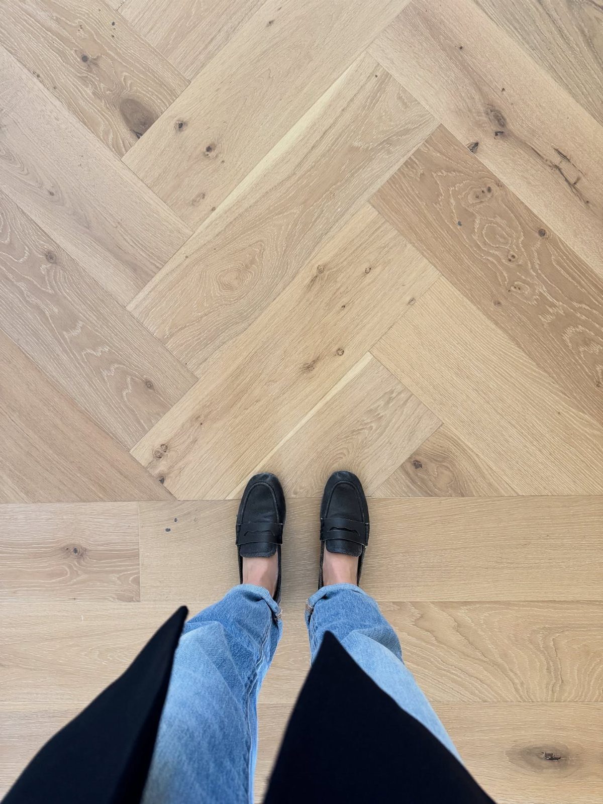 Light wood herringbone flooring with a person standing on it wearing blue jeans and black loafers.