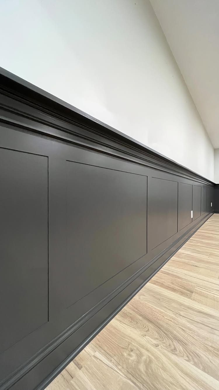Dark grey wainscoting panels installed along a white wall above light wood flooring.