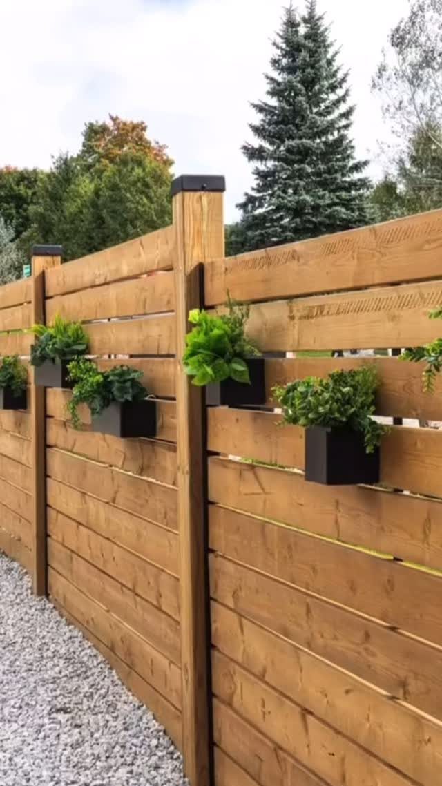 Wooden Privacy Fence: 12 Ideas That Define Your Yard 10 A light brown wooden privacy fence with black planters holding green plants along a gravel path.