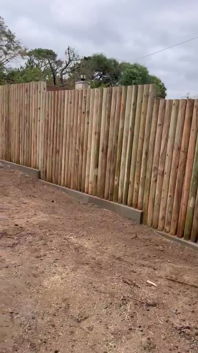 Wooden Privacy Fence: 12 Ideas That Define Your Yard 12 Vertical round timber poles forming a long wooden privacy fence along a dirt path.